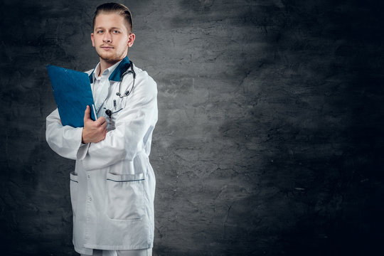 Studio portrait of young medical doctor holds blue clipboard on grey background.