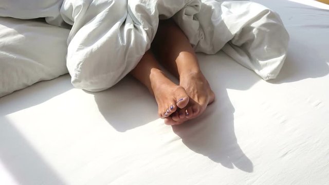 closeup of bare feet of woman in bed on a sunny morning with the blanket being pulled
