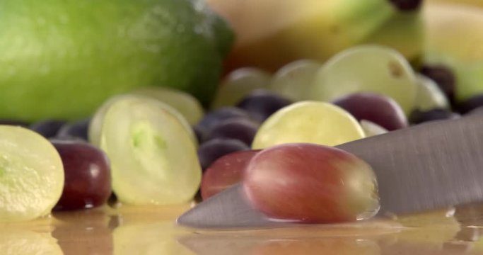 Slicing a grape in half using a sharp kitchen knife. Filmed with selection of fruit in background and on a wooden chopping board. Filmed in slow motion.