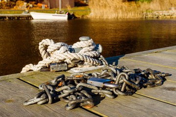 Naklejka premium Steel chain with padlock on wooden pier. Rope and padlock on bollard. Blurred background with moored boat and coastline.