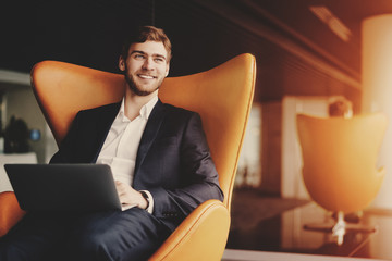 Young smiling successful man entrepreneur in formal business suite with a beard sitting on orange armchair with laptop in luxury office interior - obrazy, fototapety, plakaty