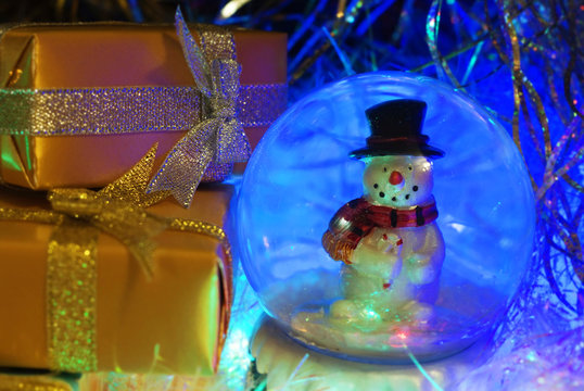 Christmas and New Year`s snowman in colorful blue glow light and gold gift boxes wrapping paper with bows of ribbons.  Greeting card blue Background with holiday tinsel.