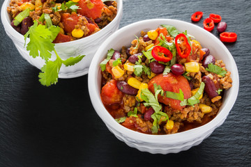 Chili con carne in a white ceramic bowl on black stone background. Cooked with ground beef, tomatoes, peppers, beans, corn, garlic, onion, cilantro.