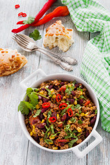Chili con carne in a white casserole on wooden table with fresh vegetables and bread. Cooked with ground beef, tomatoes, peppers, beans, corn, garlic, onion, cilantro.