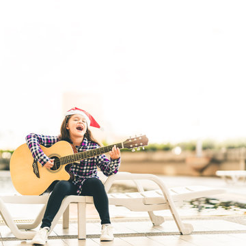 Young mixed race girl playing guitar, singing and smiling joyfully by swimming pool, with christmas santa hat, music or happiness concept, sunset warm light tone effect, with copy space
