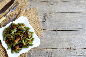Spicy fried green beans with champignons on a plate and on old wooden background with copy empty space for text. Warm green beans and champignons salad recipe. Vintage style. Top view