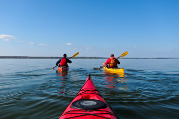 Two guys in a kayak. Kayaking in the calm blue lake. View from the red kayak. - obrazy, fototapety, plakaty