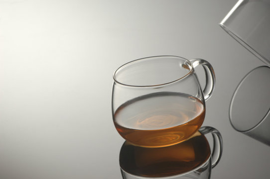 A transparent glass cup with tea on a reflecting surface and a light gray background