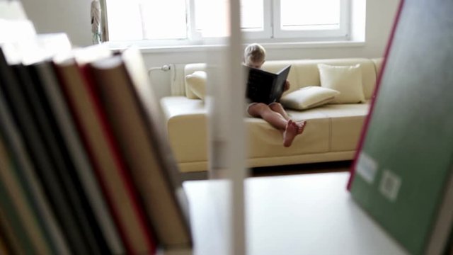 A camera peeks in on a little boy reading a book from behind a book shelf