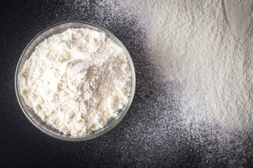 Glass bowl with a flour, on a layer of flour. On a black background.Top view. Flat lay.