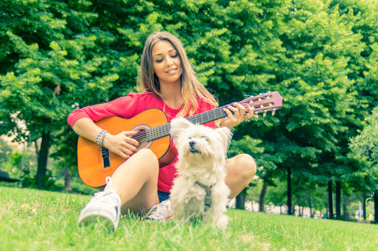 Young pretty girl playing guitar at the park with her dog. 