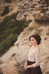 Portrait of a beautiful young caucasian stylish woman in evening gown standing on the background of spectacular nature, rock and trees, in sunlight