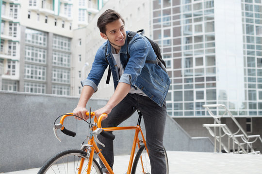 The guy goes to town on a bicycle in  blue jeans jacket .  young man  an orange fix bike
