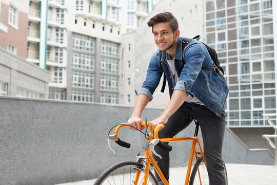 The guy goes to town on a bicycle in  blue jeans jacket .  young man  an orange fix bike