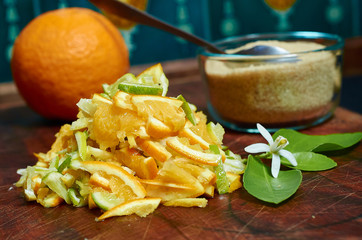 An orange, orange blossom and a pile of sliced oranges, lemons and limes on a wooden board with a bowl of sugar and a spoon.