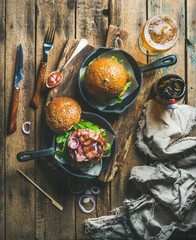 Homemade beef burgers with crispy bacon and vegetables in small cast iron pans and glass of wheat beer on rustic board over shabby wooden background, top view, vertical composition