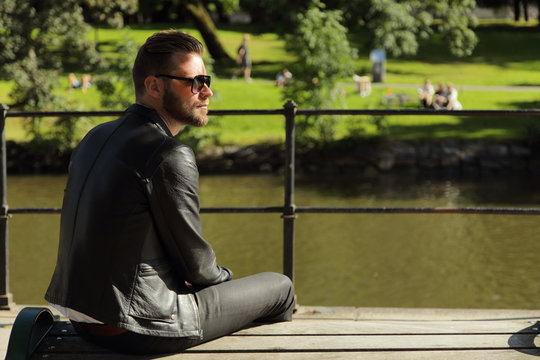 An from the side angle photo of a man sitting on a bench wearing a black leather jacket, with a river and grass behind him. Sunny summer day.