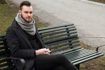 Fototapeta premium A lonely depressed man sitting down outdoors on a bench on a cold fall day wearing a black jacket, scarf and jeans.