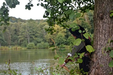 Mittelalterlicher Bogenschütze mit schwarzer Kapuze steht versteckt hinter Baum am See
