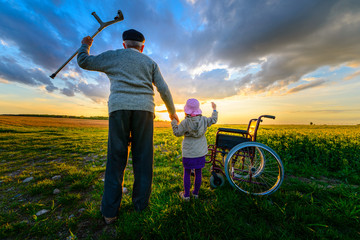 Miracle recovery: Old man gets up from wheelchair and raises hands up. Overjoyed grandfather standing up and holding hand of his granddaughter. Shot in a meadow. Recovery concept