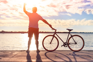 Happy man silhouette raising fist at sunset and holding sport bike - Young successful guy standing...