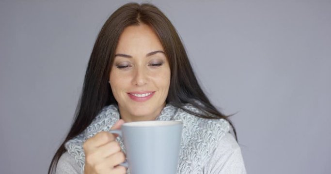 Beautiful long haired, brunette woman holding in her hand cup of hot drink. She wearing woolen scarf around her neck. She trying to warm up herself. Isolated on gray background.