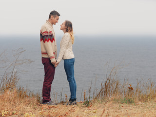 Young attractive couple in knitted sweaters standing on a cliff at sea shore and holding hands
