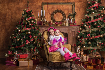 Fototapeta na wymiar Cute sisters sitting on armchair near christmas trees, wearing pink skirts and red headbands. Smiling toddlers. Christmas gifts.