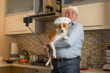 Funny mature man doing selfie in a kitchen with cute basenji dog wearing white cap