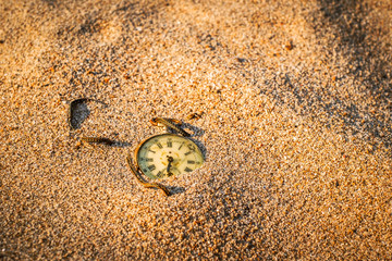 Old pocket watch with cracked glass partially buried in sand on the beach at the sunset