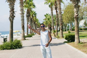 A nice guy in white suit standing in a park with palm trees and showing two fingers