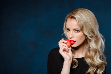 Beautiful blonde girl in black dress holding apple, looking at camera.