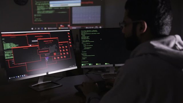 Male hacker hacks computer in dark. Computer code reflecting on his face. HD.
