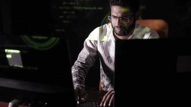 Male hacker hacks computer in dark. Computer code reflecting on his face. HD.