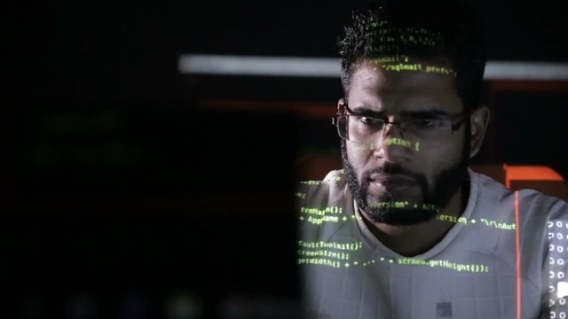Male hacker hacks computer in dark. Computer code reflecting on his face. HD.