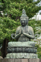 Fototapeta na wymiar Tokyo, Japan - September 26, 2016: Closeup of bronze statue of Kannon Bosatsu, attendant of Amida Buddha, sitting on lotus pedestal in garden at Senso-ji Buddhist Temple. Green foliage. 