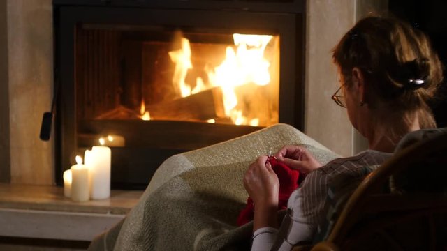 Grandmother knits a sweater sitting in front of a fireplace