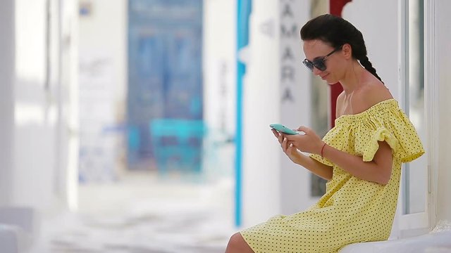 Beautiful woman with smart phone outdoors in old streets an Mykonos. Girl at street of typical greek traditional village with white walls and colorful doors on Mykonos Island, in Greece, Europe