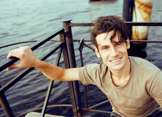 young pretty smiling hipster guy on river dock, traveling lifestyle modern people concept