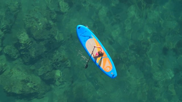 AERIAL: Girl rider standup paddleboarding in beautiful crystal clear emerald sea