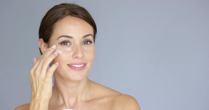 Beautiful young woman applying face cream to her cheek bone in a beauty  skincare or cosmetics concept  on grey with copy space