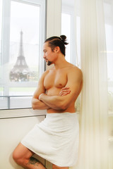young man in white towel indoor looking out of window in the morning with Eiffel tower in Paris, France on the background, soft focus. Black and white vintage style picture.