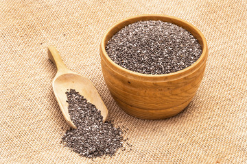 Wooden bowl full with chia seeds and a spoon next to it on sack cloth background seen from above