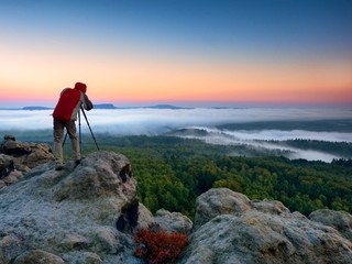 Photographer takes photos with mirror camera and tripod on peak of rock. Fall fogy landscape, spring orange pink misty sunrise in beautiful valley below.