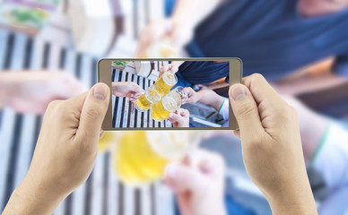 Man is using mobile phone taking photo of a group of people clink glasses of beer
