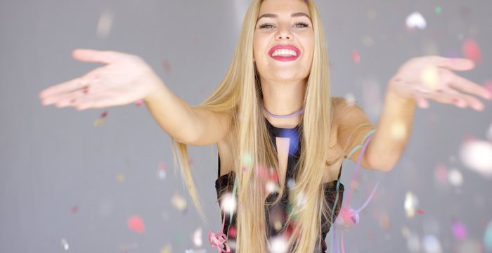 Sexy, blond girl blowing confetti from her hands to camera direction. She dancing and having fun on a new year party. Smiling to the camera.