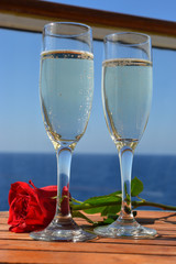 Flutes of champagne and red rose on table of cruise ship with ocean in the background.
