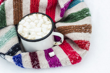 Fototapeta na wymiar Old metal cup of hot cocoa with marshmallows and a soft colorful knitted scarf on a fluffy the snow close-up