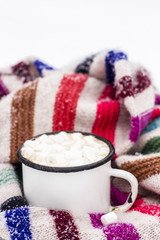 Old metal cup of hot cocoa with marshmallows and a soft colorful knitted scarf on a fluffy the snow close-up