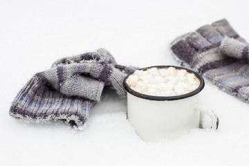 Old metal cup of hot cocoa with marshmallows standing in the snow next to lie a striped knitted the gloves